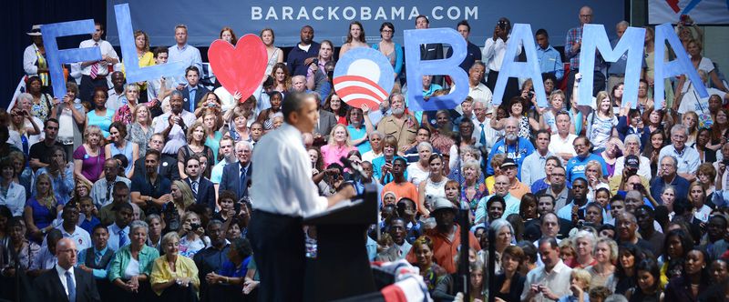 Obama, in dark slacks and a white shirt, stands at a podium surrounded by supporters, a group of whom hold aloft blue and red letters reading: “FL [heart symbol] Obama”.