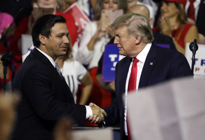 Trump, in a dark suit, white shirt, and red tie, smiles slightly as he shakes hands with a smiling DeSantis, who wears a dark suit, white shirt, and silver tie, in front of a cheering crowd.