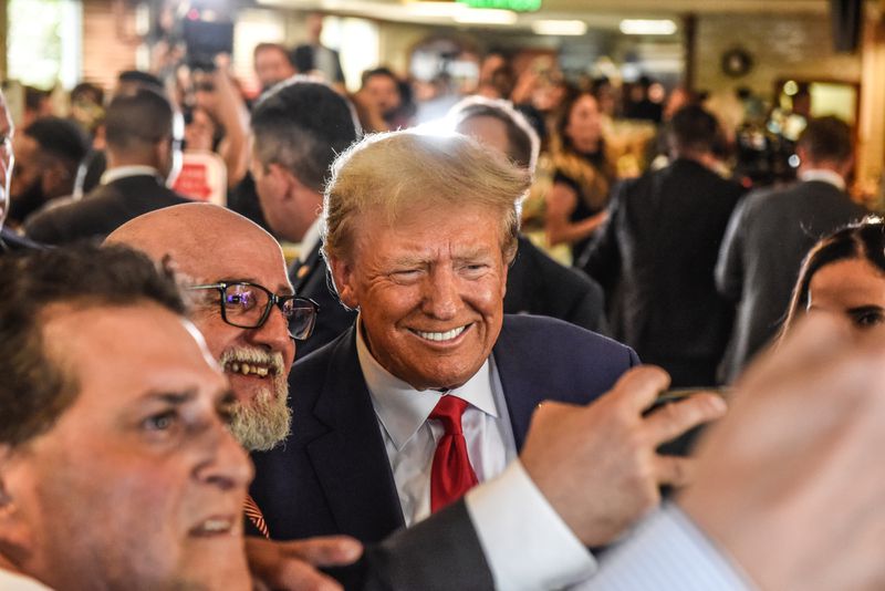 Trump, in a navy suit, white shirt, and red tie, smiles broadly as he stands close to a white bearded, bald man wearing black framed glasses, who holds his phone out for a selfie.