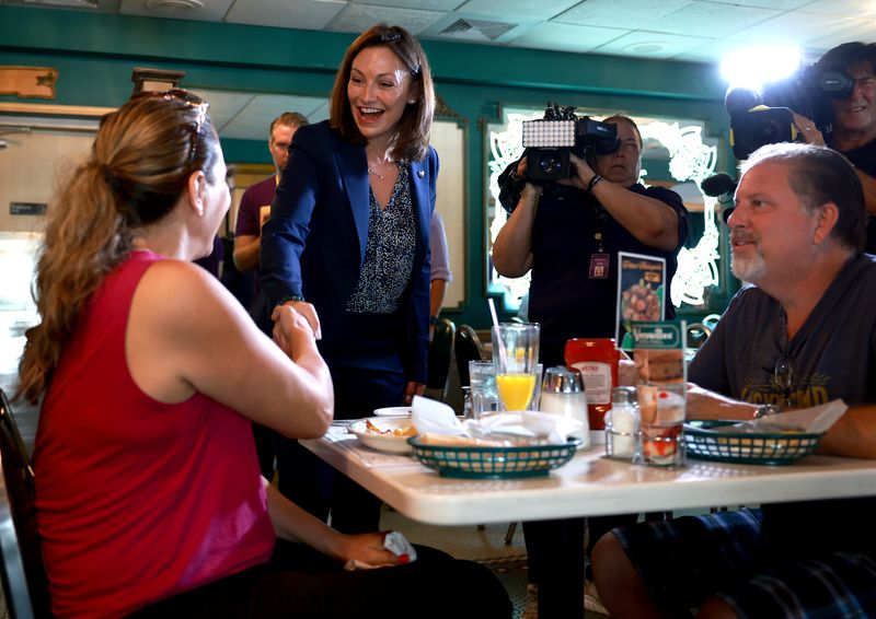 Fried, smiling brightly in a blue suit and black-and-white blouse, her brown hair in a bob, shakes hands with a ponytailed woman in pink, who is dining with with a gray-haired man in a blue T-shirt. Cameras behind her record the encounter.