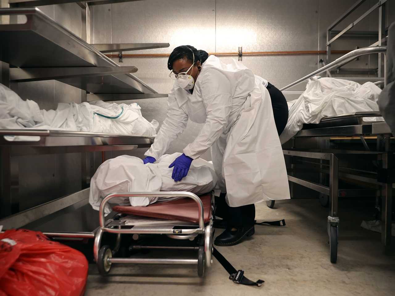 A worker wearing a mask, surgical gown, and gloves places a body wrapped in a sheet onto a stretcher inside a morgue.