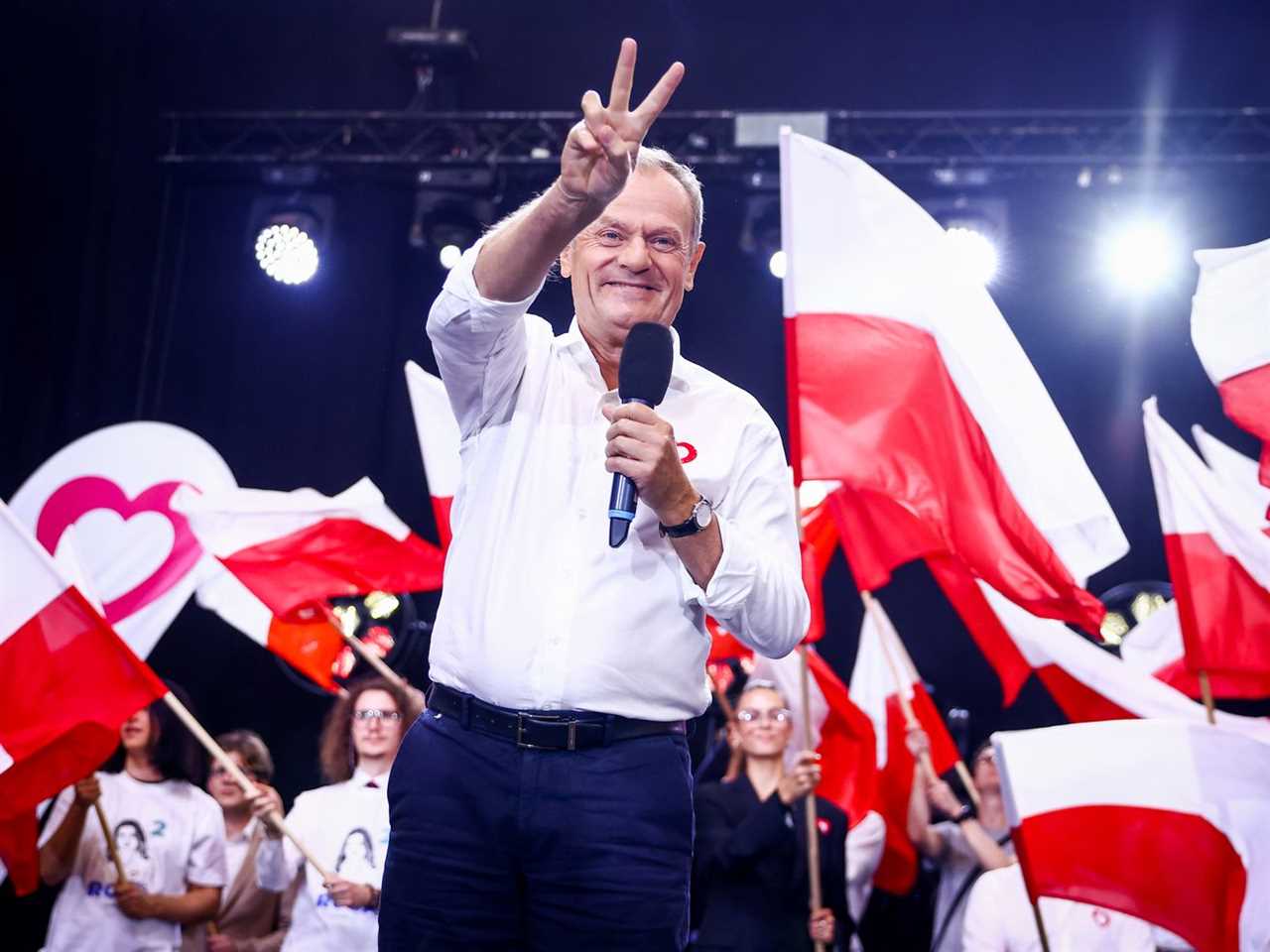 Donald Tusk, amid waving red-and-white flags, holds a microphone in one hand and makes a V-sign with the other.