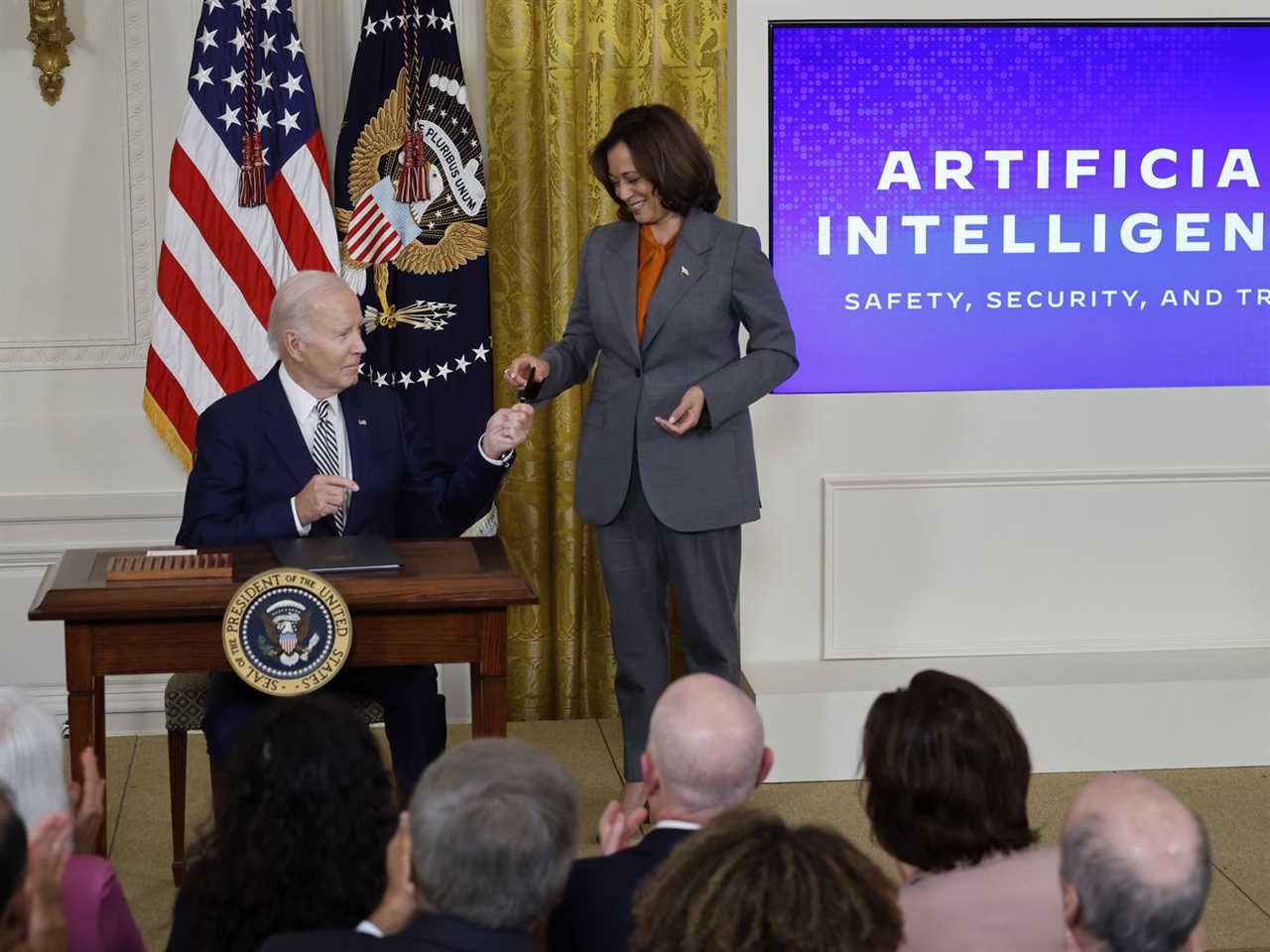 President Joe Biden, sitting at a desk, hands his pen to Vice President Kamala Harris, standing, while a presentation on AI is projected on a screen next to them.