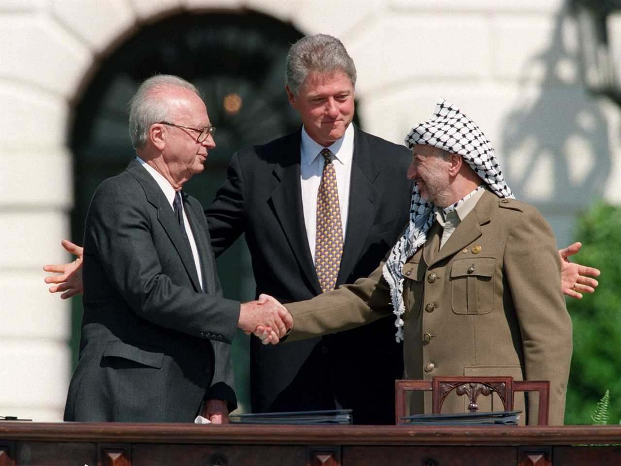 US President Bill Clinton, gray haired and clean shaven in a dark suit and black and gold tie, stands behind Rabin (white haired in a dark suit) and Arafat&nbsp;(in green fatigues and a black and white keffiyeh) as the two Middle Eastern leaders shake hands.