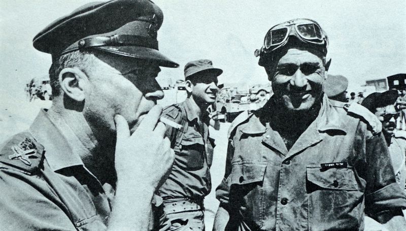 In a blue-tinted black and white photo, Rabin, close to the camera and seen in profile, smokes a cigarette. Gavish, in his fatigues, desert goggles around his military cap, smiles at the camera. Uniformed men move with purpose behind them.