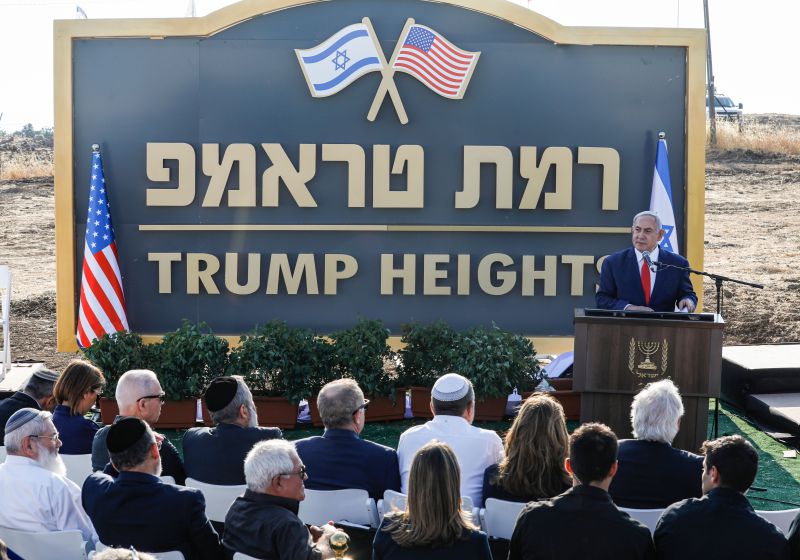 Out in the desert, before a seated crowd, Netanyahu, in a blue suit, white shirt, and red tie stands at a podium, set up in front of a large blue and gold sign featuring the US and Israeli flags. In Hebrew, the sign says Ramat Trump; below in English it reads Trump Heights.