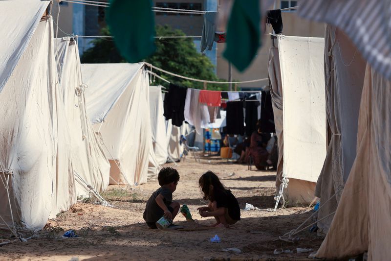 A boy and a girl, each perhaps about 3 years old, are seen from behind as they use discarded items, including a can, to dig in the dirt “street” created by a long row of white tents. Laundry hangs on lines strung over their play area.