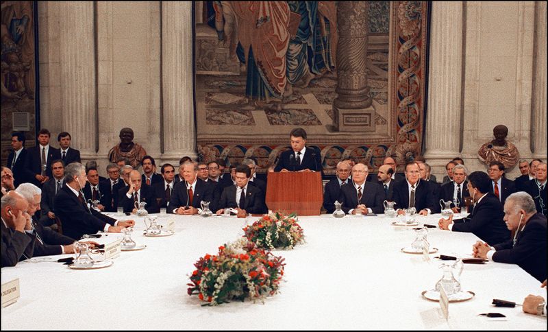 Men, many of them grey-haired, sit in dark suits around a long rectangular table with red and white flower arrangements. They are packed shoulder to shoulder, and listen with headphones in their ears to Gonzalez, also in a dark suit, speaking at a wooden lectern set atop the table. A posse of dark-suited men sit in a line behind him, also densely packed together. A tapestry that seems to feature robed figures hangs behind them.