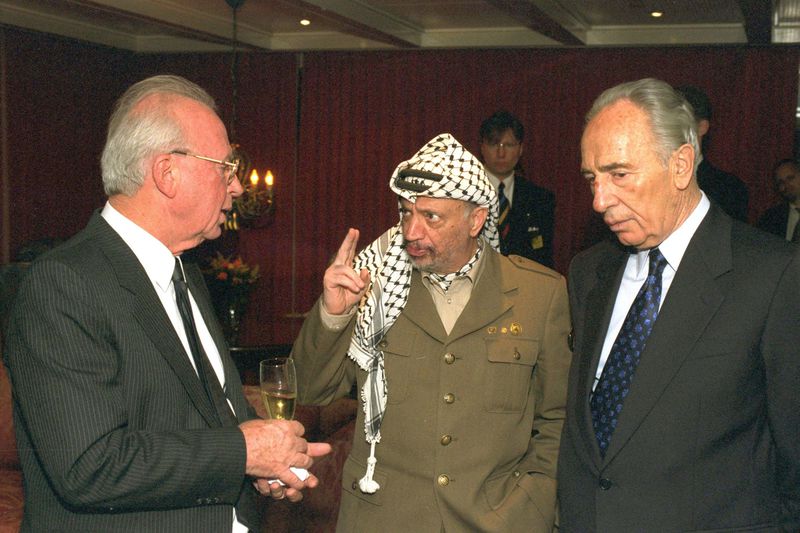 Arafat, in his military uniform and keffiyeh, stands between the Israeli men and holds up two fingers, looking serious. Rabin, in a black suit and tie, holds a glass of champagne, while Peres seems to have his hands in his pockets. Both men listen intently.