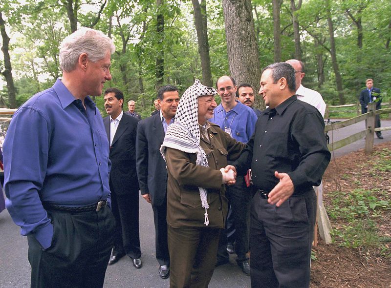 Clinton, white-haired and clean shaven, in a loose-fitting blue dress shirt and dark slacks, smiles as he watches Arafat, dressed in his military uniform and keffiyeh, shake hard with Barak, dressed in a black dress shirt and black slacks. Their aides mill about behind them and the whole group is surrounded by trees fulll of green leaves.