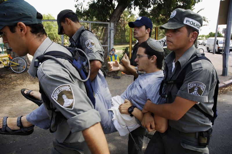 Four officers carry a young man with glasses, dark hair, and a yarmulke between them, each holding one of his limbs, transporting him down a dusty road. He wears an expression that appears angry and defiant.