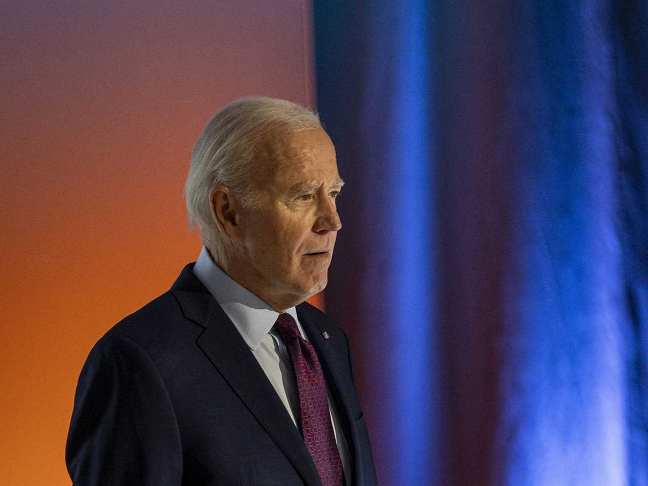 US President Joe Biden standing in front of a blue curtain and orange background.