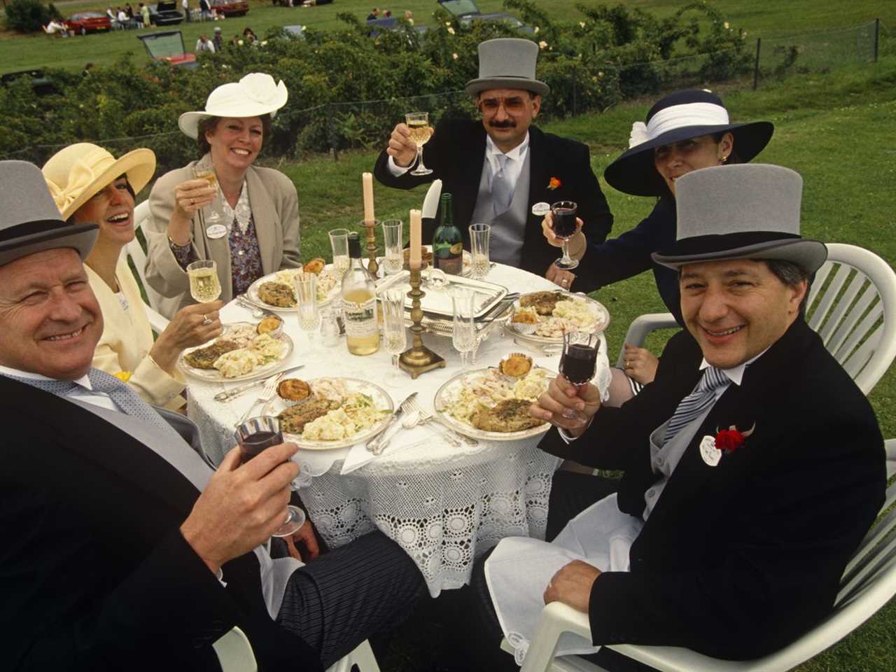 Three fancily dressed English couples sit around a white-clothed round table, at a formal picnic lunch for Royal Ascot racegoers on Ladies’ Day. 