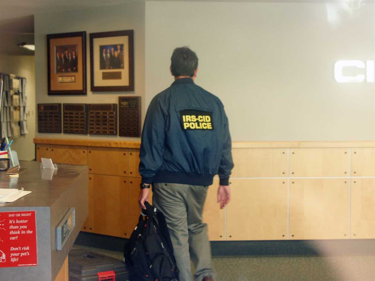 A person wearing an IRS police jacket walks through a house.