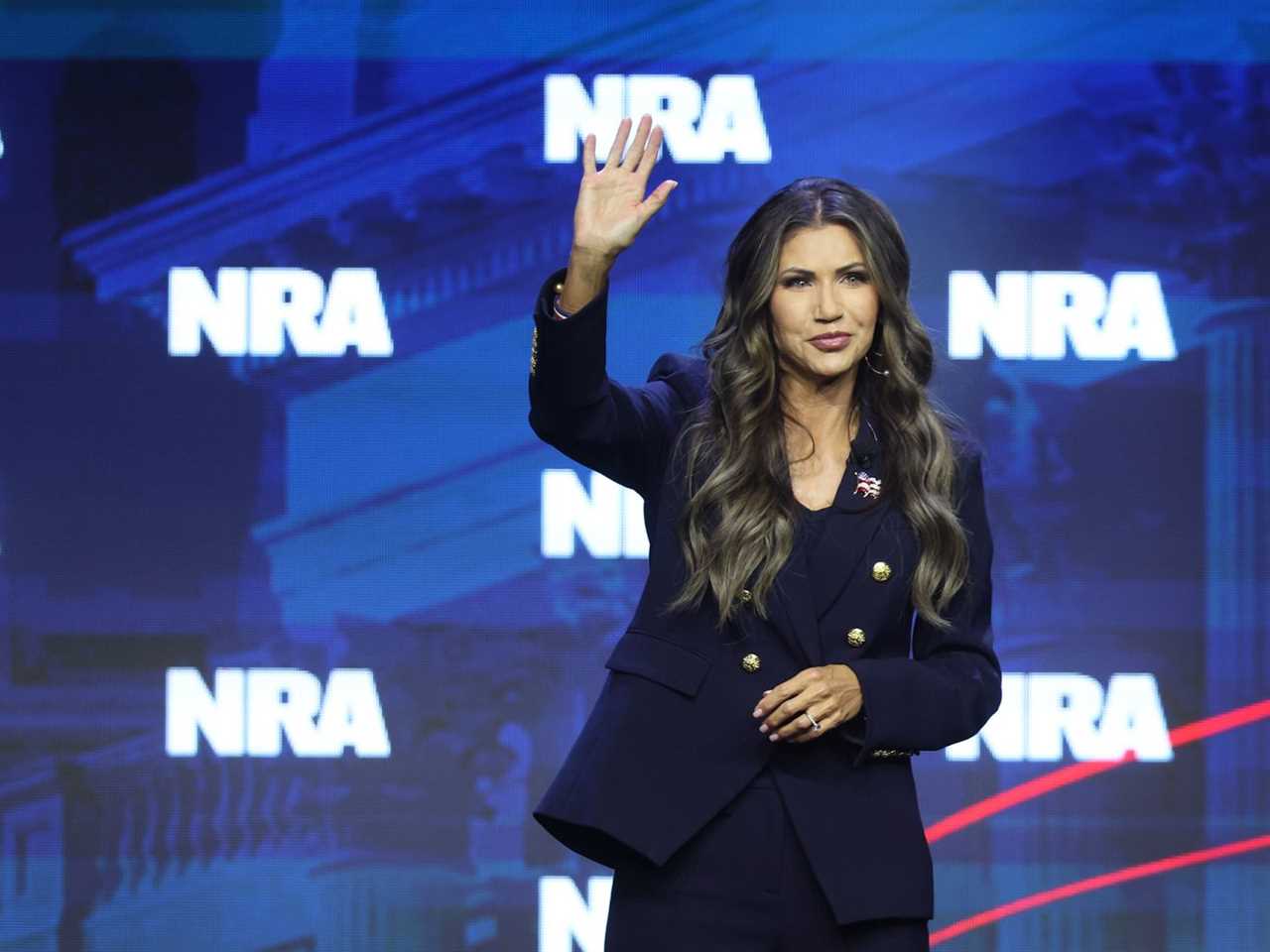 Kristi Norm waves to the audience from a stage dressed with an “NRA” backdrop.