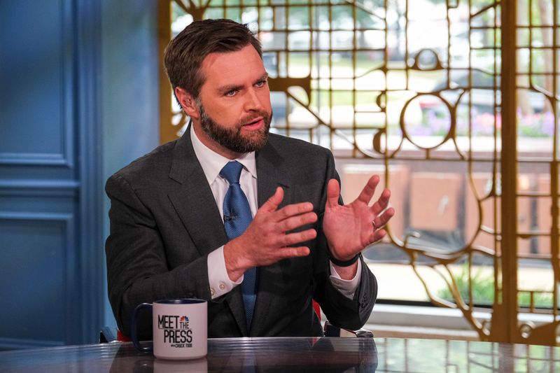 Vance, mid-conversation, gestures with his hands. He wears a blue tie and gray suit. He sits in front of an ornately designed window. On the table in front of him sits a coffee cup that reads “Meet the Press.”