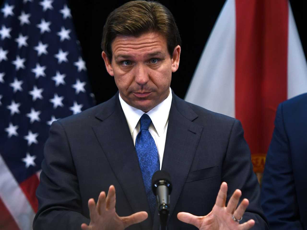 DeSantis gestures with both hands while holding a press conference, standing in front of a US flag.