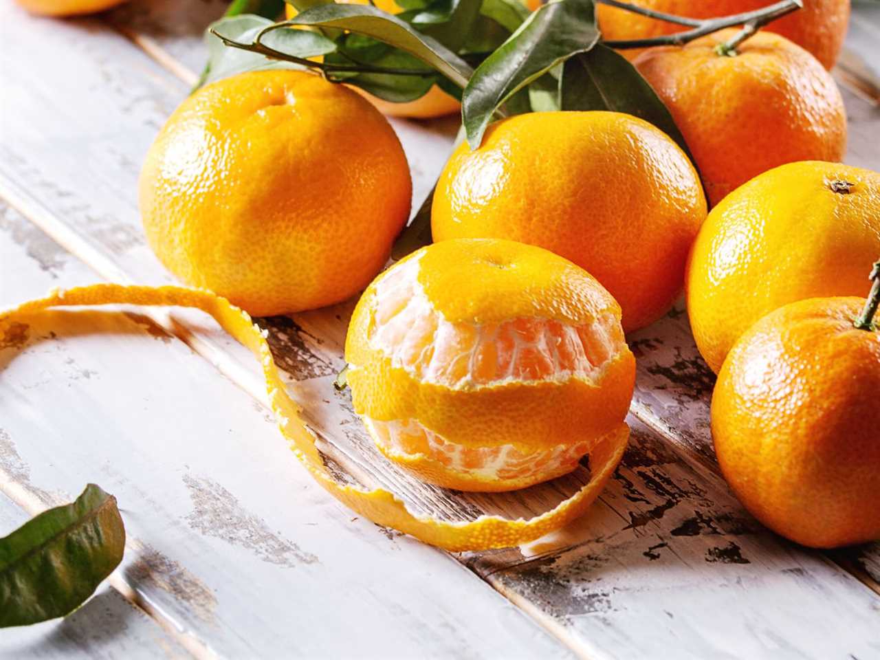 Ripe orange clementines or tangerines with leaves sit on a white wooden plank table, with one in front partially peeled.