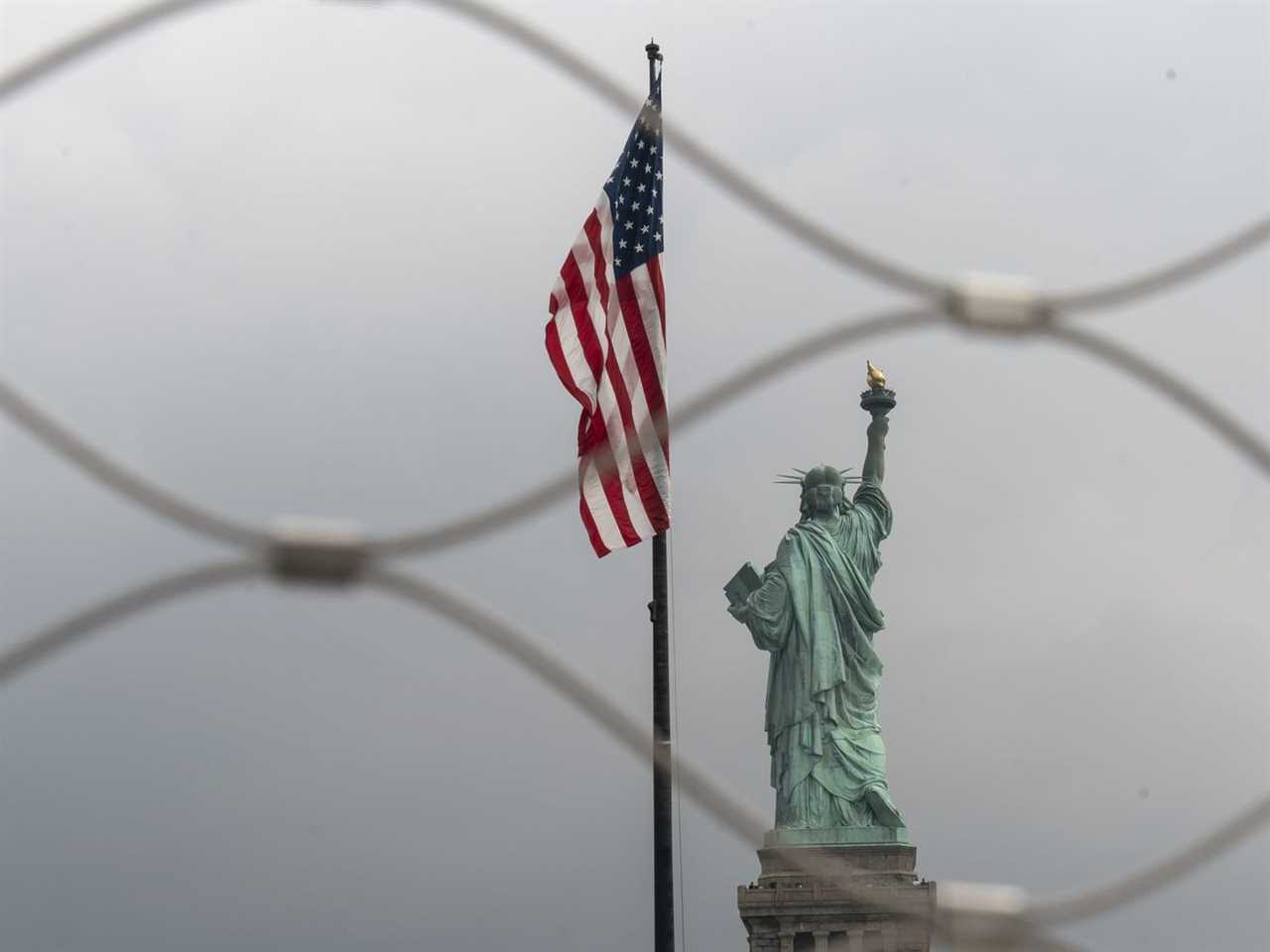 The Statue of Liberty and an American flag, photographed through wire fencing.
