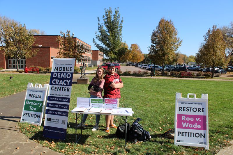 Two people stand back-to-back behind a white folding table, set up near signs that read, “MObile democracy center” and “Restore Roe v. Wade petition.”