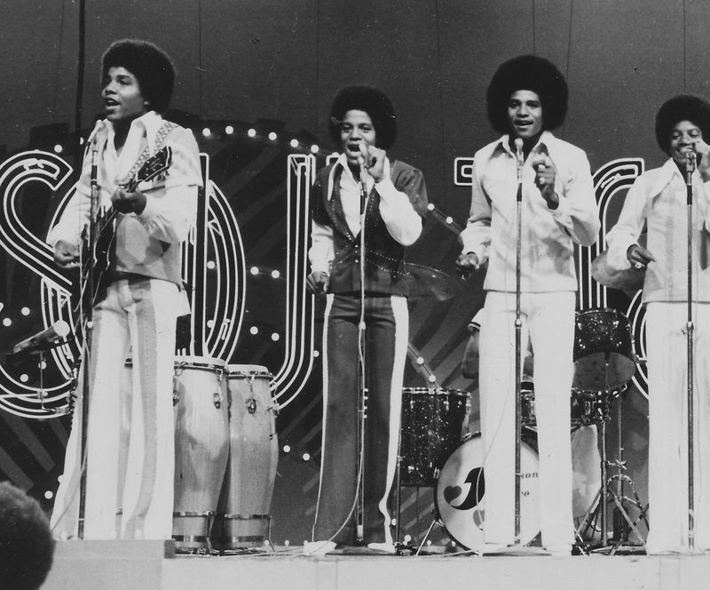 A black and white photo of the Jackson Five singing and playing guitar on the Soul Train stage. They are wearing black and white suits and have afros.