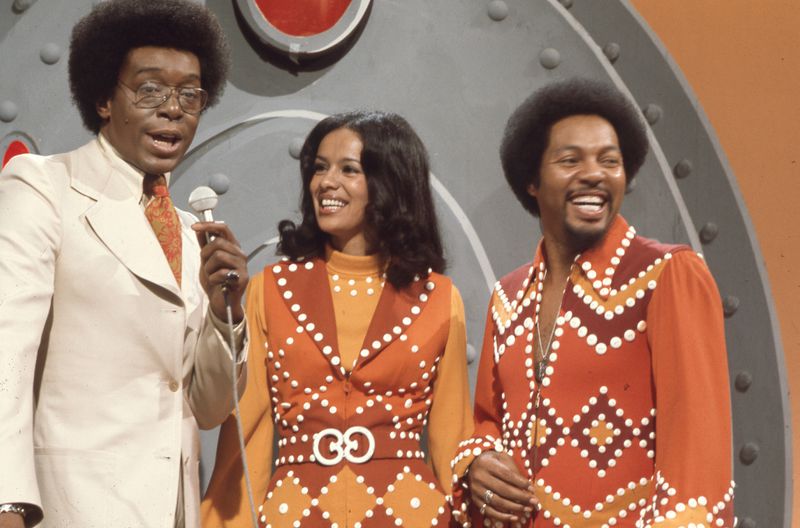 Don Cornelius is wearing a khaki suit and speaking into a microphone. Next to him Marilyn McCoo and Billy Davis Jr. are wearing matching brightly-colored outfits with white dot details. They’re laughing.