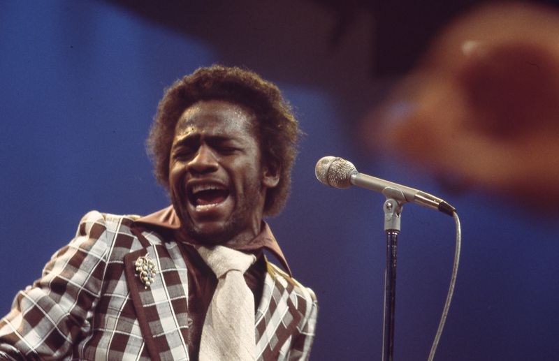 Al green singing loudly into a microphone with his eyes closed. He’s wearing a bold, plaid suit with a wide tie.