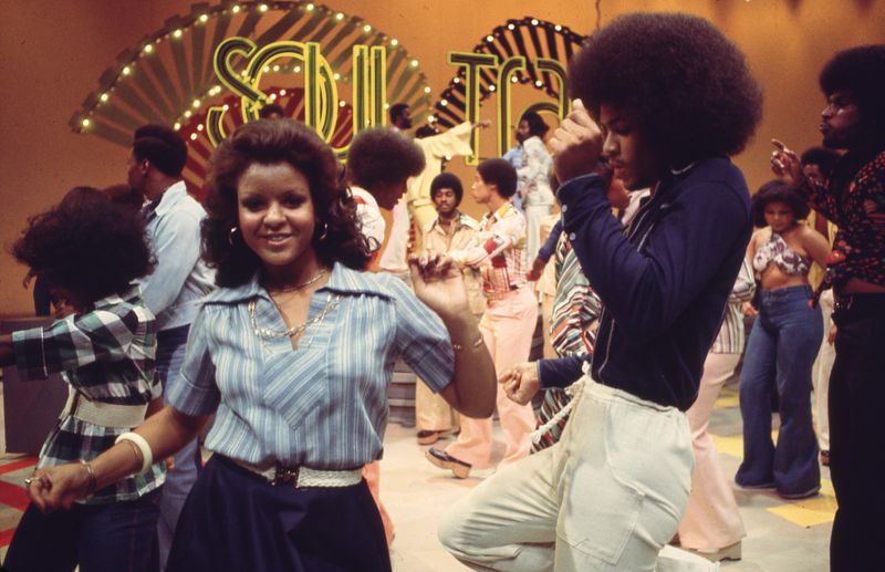 People wearing bright, layered 70s-style clothing are dancing in front of the Soul Train stage.