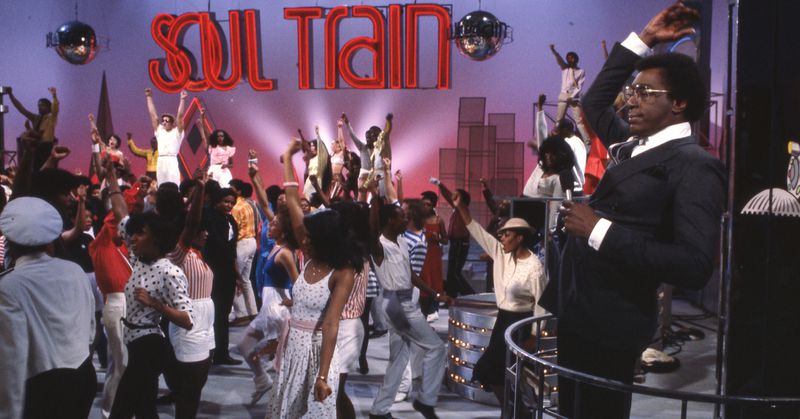 Don Cornelius stands holding a microphone and raising a hand. The Soul Train Dancers are dancing between him and the performers on stage.