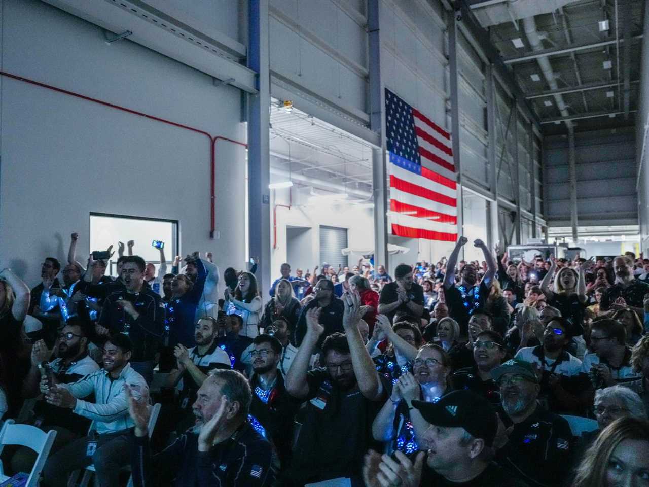 A hangar-sized room with an American flag on one wall and a crowd of cheering people.