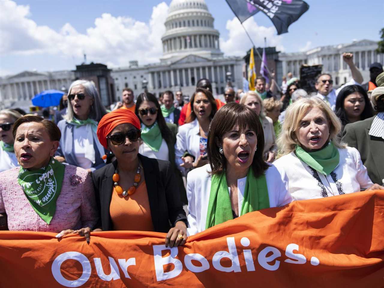 Rows of women march in front of the US Capitol building, the front row carrying an orange banner.