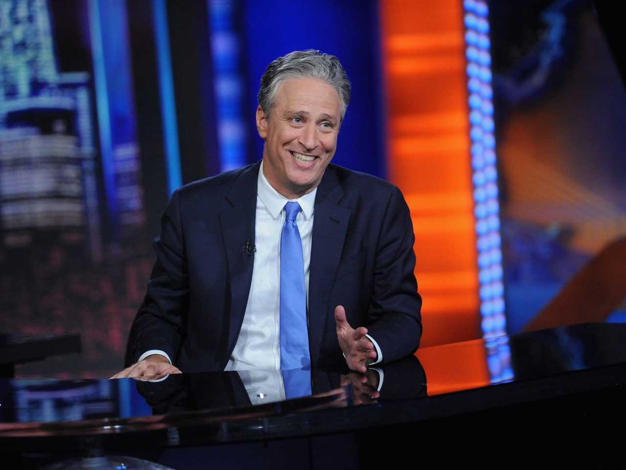 Jon Stewart sits at a desk wearing a black suit jacket and light blue tie, smiling. 