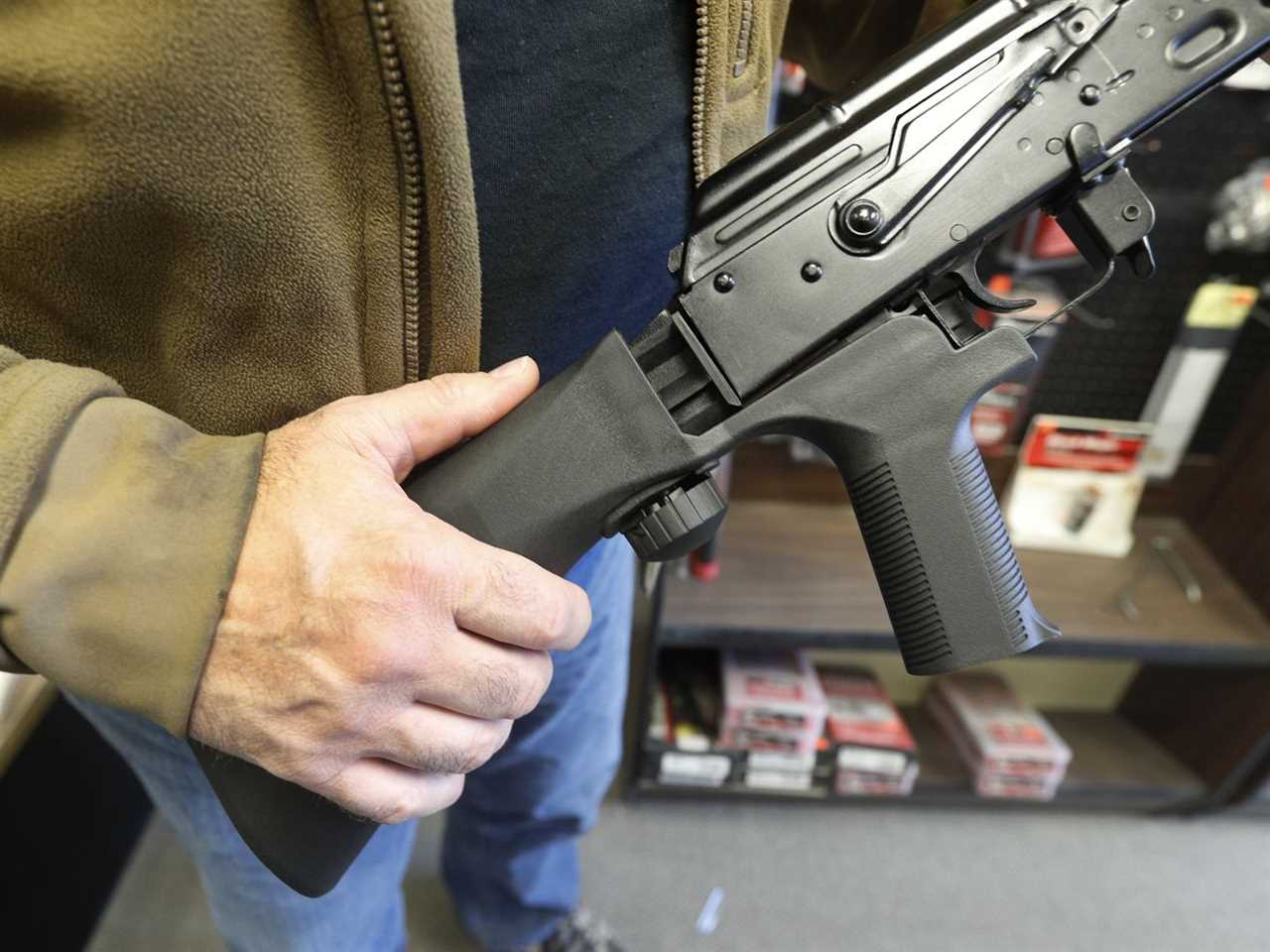 A man wearing a brown jacket and jeans holds a bump stock on the left while fitting it onto an AK-47 semi-automatic rifle on the right. Behind him are shelves of ammunition.