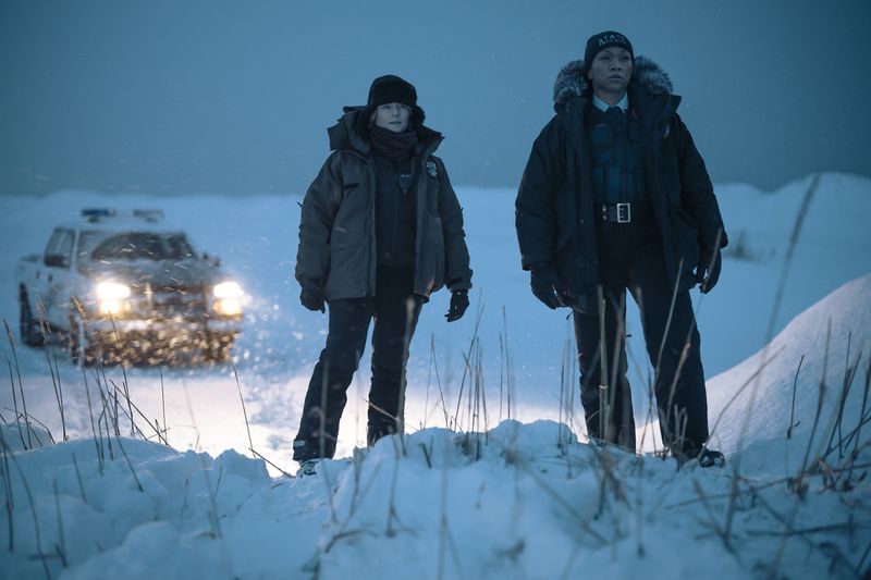 Two women in police uniforms and winter gear look out over a snowy landscape, a police SUV with its lights on illuminating the scene.