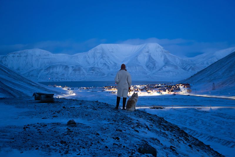 A woman wearing a coat stands with a dog on a hill within a dark blue landscape. They are looking down at a small town lit by the lights on the buildings.