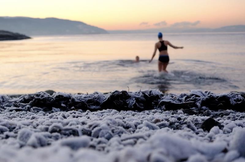 A woman in a swimsuit and swim cap is wading into a body of water with another person. There is low, warm light and snow on the ground.