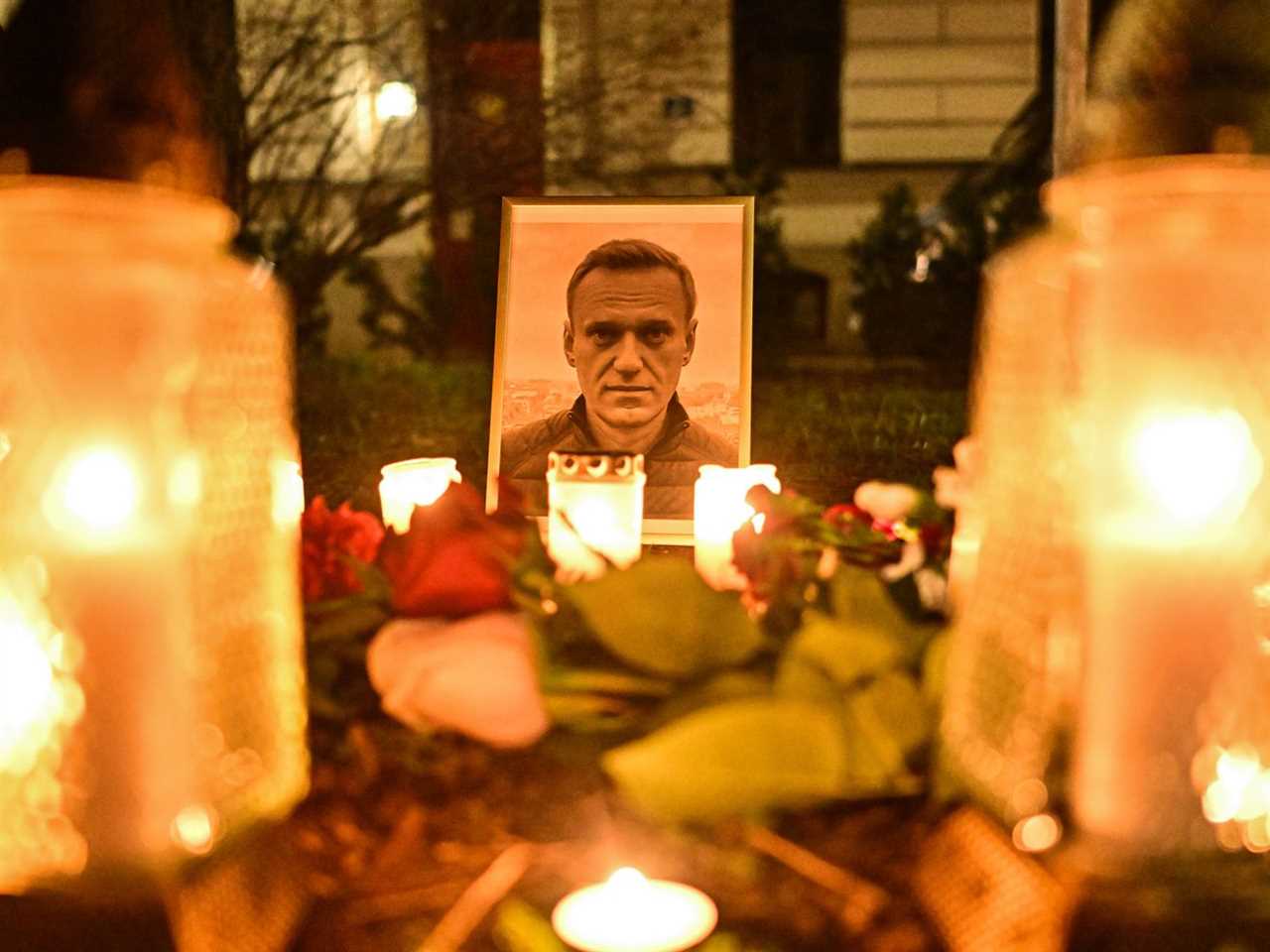 A photo of a row of yellow candles in glass jars; at the end surrounded by roses, is a color photo of Navalny, a white clean shaven man with sandy brown hair.