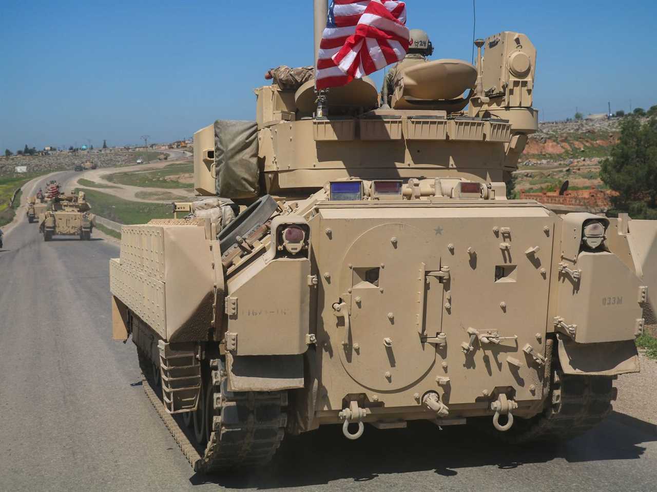 An armored vehicle flying an American flag traveling down a desert road.